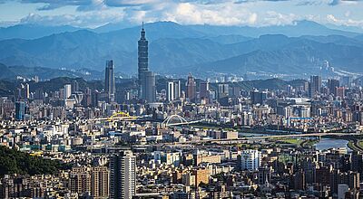 Panoramaaufnahme von Taipeh, Taiwan, mit dem markanten Taipei 101 Wolkenkratzer im Zentrum, umgeben von zahlreichen modernen Hochhäusern. Im Vordergrund ist ein dichter Stadtbereich mit Wohn- und Geschäftsgebäuden zu sehen, im Hintergrund erheben sich bewaldete Berge unter einem teils bewölkten Himmel. Zwei auffällige Brücken überspannen einen Fluss, der sich durch die Stadt schlängelt.
