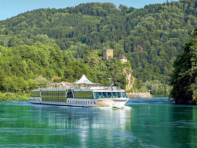 Ein weißes Flusskreuzfahrtschiff passiert bei Sonne einen Berg mit dichten grünen Laubbäumen