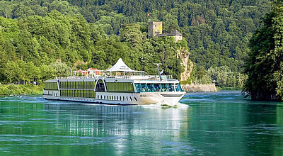 Bereits im April soll die Arielle Royal erste Donau-Kreuzfahrten unter dem Label Arielle Kreuzfahrten unternehmen. Modell: Reisen Aktuell Ein weißes Flusskreuzfahrtschiff passiert bei Sonne einen Berg mit dichten grünen Laubbäumen