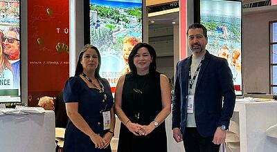 BU: Von links nach rechts: Nifüfer Kati (Sales Direktorin), Nilda Türe (Sales Koordinatorin), Ilhan Yalcin (Sales Direktor). Zwei Frauen und ein Mann stehen lächeln vor einigen Leuchtreklamen und Stehtischen und lächeln in die Kamera.