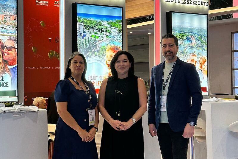 Zwei Frauen und ein Mann stehen lächeln vor einigen Leuchtreklamen und Stehtischen und lächeln in die Kamera.