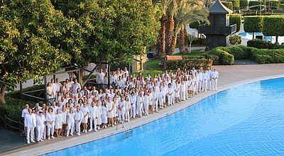 Die Teilnehmer der Amondo-Tagung im Limak Lara Deluxe Hotel Große Gruppe von Menschen in weißer Kleidung steht am Rand eines Hotelpools in einer grünen, palmenreichen Ferienanlage bei Sonnenuntergang und blickt lächelnd in Richtung Kamera.