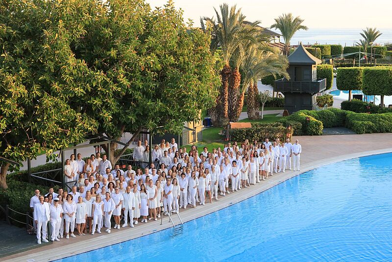 Die Teilnehmer der Amondo-Tagung im Limak Lara Deluxe Hotel Große Gruppe von Menschen in weißer Kleidung steht am Rand eines Hotelpools in einer grünen, palmenreichen Ferienanlage bei Sonnenuntergang und blickt lächelnd in Richtung Kamera.