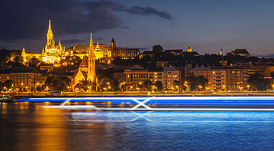 Das Markenzeichen „X“ soll ab 2027 auch auf Flüssen zu sehen sein Ein stark stilisiertes, futuristisches Flusskreuzfahrtschiff aus leuchtendblauen Streifen und mit Markenzeichen X fährt im Abendlicht auf einem Fluss. Im Hintergrund sind erleuchtete Häuser und Kirchen zu sehen