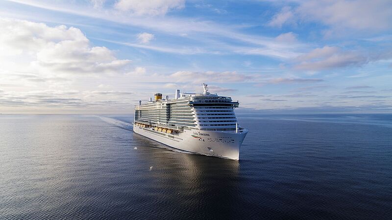 Kreuzfahrtschiff Costa Toscana fährt bei ruhiger See unter blauem Himmel über das offene Meer, aufgenommen aus der Luft von vorne links. Das moderne Schiff zeigt mehrere Decks mit Balkonkabinen und markantem gelbem Schornstein.