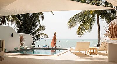 Frau in Sommerkleid und Hut steht am Rand eines Infinity-Pools mit Blick auf das Meer, umgeben von Palmen und moderner, offener Architektur in einem luxuriösen Strandresort.