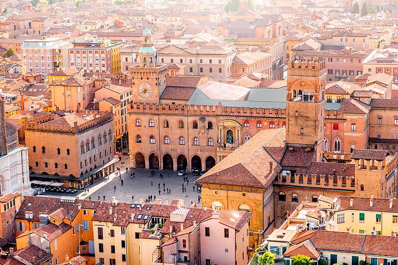 Luftaufnahme des zentralen Platzes Piazza Maggiore in Bologna, Italien. Im Mittelpunkt steht der historische Palazzo d'Accursio mit Uhrturm, flankiert von weiteren mittelalterlichen Gebäuden mit roten Ziegeldächern. Menschen spazieren über den Platz, der von farbenfrohen Häusern und engen Gassen umgeben ist. Die Szene ist in warmes Sonnenlicht getaucht und vermittelt eine lebendige Atmosphäre.