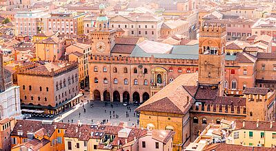 Luftaufnahme des zentralen Platzes Piazza Maggiore in Bologna, Italien. Im Mittelpunkt steht der historische Palazzo d'Accursio mit Uhrturm, flankiert von weiteren mittelalterlichen Gebäuden mit roten Ziegeldächern. Menschen spazieren über den Platz, der von farbenfrohen Häusern und engen Gassen umgeben ist. Die Szene ist in warmes Sonnenlicht getaucht und vermittelt eine lebendige Atmosphäre.