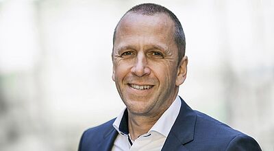 Ein Mann mittleren Alters lächelt in die Kamera. Der Mann trägt einen Anzug und ein weißes Hemd.