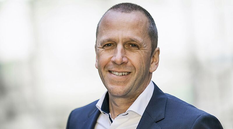 Ein Mann mittleren Alters lächelt in die Kamera. Der Mann trägt einen Anzug und ein weißes Hemd.