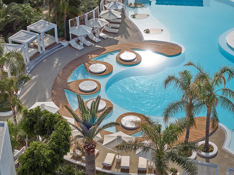 Das Caramel wird im kommenden Jahr als erstes All-Inclusive-Boutique-Resort der Gruppe neu positioniert. Luxuriöse Poolanlage in einem Resort mit geschwungenen Holzdecks, runden Loungeplattformen im Wasser, weißen Liegen und Sonnenschirmen sowie umgebenden Palmen und üppiger Vegetation, fotografiert aus der Vogelperspektive.