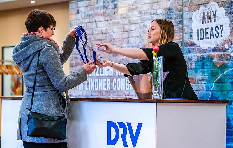 Eine Frau im grauen Mantel erhält am Empfangstresen von einer Mitarbeiterin ein Namensschild mit blauem Lanyard. Auf dem Tresen steht eine Vase mit einer roten Tulpe, im Hintergrund ist eine Backsteinwand mit der Aufschrift ‚ANY IDEAS?‘ zu sehen. Auf der Theke prangt das Logo ‚DRV‘.