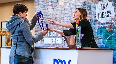 Eine Frau im grauen Mantel erhält am Empfangstresen von einer Mitarbeiterin ein Namensschild mit blauem Lanyard. Auf dem Tresen steht eine Vase mit einer roten Tulpe, im Hintergrund ist eine Backsteinwand mit der Aufschrift ‚ANY IDEAS?‘ zu sehen. Auf der Theke prangt das Logo ‚DRV‘.