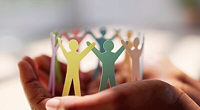 Eine Nahaufnahme von zwei Händen, die eine bunte Papierkette aus stilisierten, menschlichen Figuren in einem Kreis halten. Die Figuren sind in verschiedenen Farben wie Gelb, Grün, Rosa und Lila gehalten und halten sich an den Händen, was Gemeinschaft und Zusammenhalt symbolisiert. Der Hintergrund ist weich und unscharf, was den Fokus auf die Papierfiguren und die schützenden Hände legt.