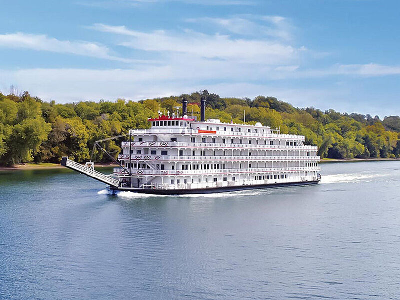 Der Schaufelraddampfer American Splendor ist auf 180 Gäste ausgelegt. Foto: Nicko Cruises Ein weißes, hohes Schaufelradschiff fährt auf dem blauen Mississippi, am Ufer stehen grüne Bäume, darüber ist hellblauer Himmel zu sehen