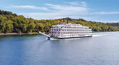 Ein weißes, hohes Schaufelradschiff fährt auf dem blauen Mississippi, am Ufer stehen grüne Bäume, darüber ist hellblauer Himmel zu sehen