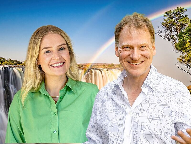Ein lächelnder Mann und eine lächelnde Frau stehen nebeneinander vor dem Hintergrund eines großen Wasserfalls mit Regenbogen. Die Frau trägt ein grünes Hemd, der Mann ein weißes Hemd mit geometrischem Muster. Der Himmel ist blau und die Szene wirkt hell und freundlich.