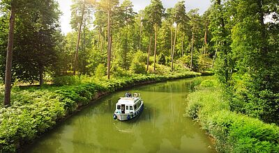 Ein weißes Hausboot unterwegs auf einem ruhigen Fluss mit grünem Wasser, am Ufer wachsen Farne und hohe Bäume.