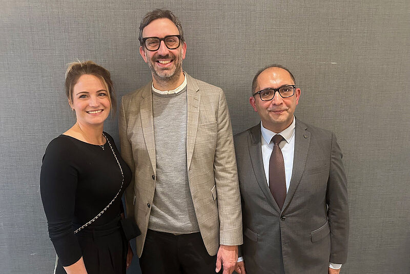 Drei lächelnde Personen stehen vor einer grauen Wand