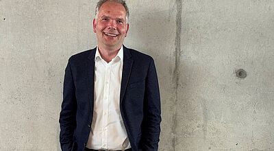 Lächelnder Mann mit weißem Hemd, dunklem Jackett und beiger Hose steht vor einer Betonwand