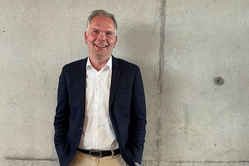 Lächelnder Mann mit weißem Hemd, dunklem Jackett und beiger Hose steht vor einer Betonwand