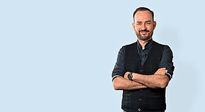 Inzwischen ist Marabu als Ferienflieger in der Branche angekommen, meint CEO Axel Schefe Professionelles Porträt eines Managers in Business-Casual-Kleidung mit schwarzer Weste und kariertem Hemd vor hellblauem Hintergrund.