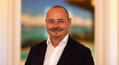 Gewährt Reisebüros zehn Prozent Provision ab der ersten Buchung: Dirk Gowin, Geschäftsführer von Select Luxury Travel Porträt eines lächelnden Mannes mittleren Alters mit kurzem Haar und Bart, der in einem dunklen Sakko und weißem Hemd vor einem unscharfen, farbenfrohen Hintergrund steht.
