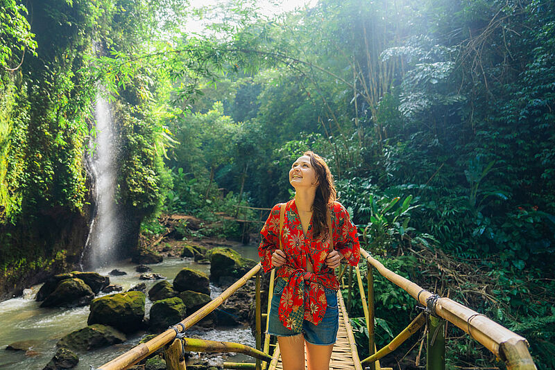 Junge Frau in roter Bluse auf Bohlenweg aus Bambus vor Wasserfall auf Bali