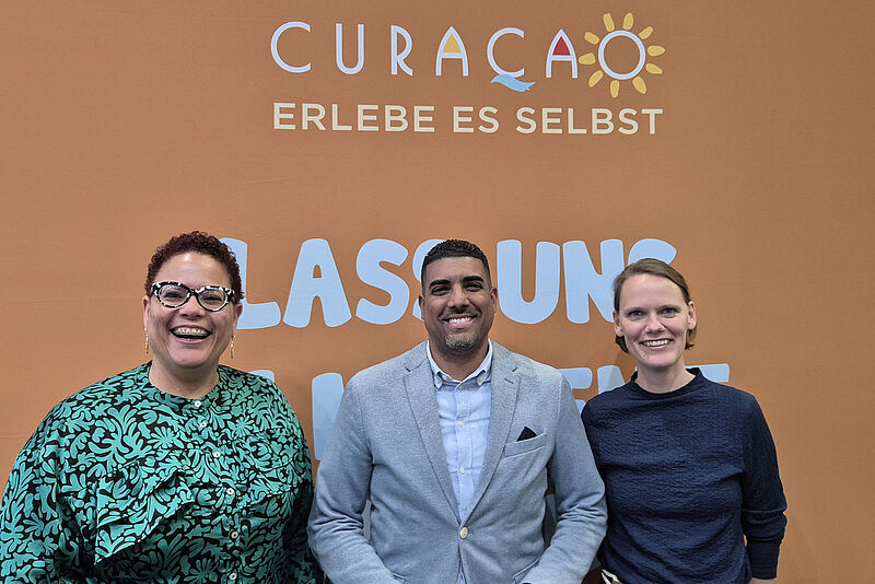Drei lachende Personen unter einem Plakat mit Curacao-Schriftzug