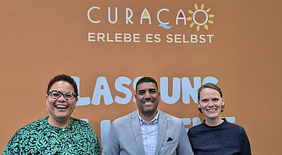 Drei lachende Personen unter einem Plakat mit Curacao-Schriftzug