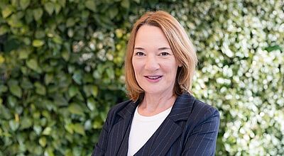 Heike Niederberghaus freut sich, mit dem neuen Feature Reisebüros einen „ausdrücklichen Wunsch“ erfüllen zu können. Foto: Explorer Porträt einer selbstbewussten Frau mittleren Alters mit schulterlangem, hellbraunem Haar, die in einem dunkelblauen Nadelstreifen-Blazer und weißem Shirt vor einer grünen Pflanzenwand steht. Sie blickt freundlich in die Kamera und hat die Arme verschränkt.