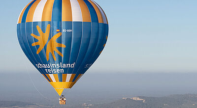 Der neue Heißluftballon von Schauinsland-Reisen wird künftig auch für touristische Fahrten genutzt Heißluftballon von Schauinsland-Reisen in blau-gelb mit Registrierung EC-OOH schwebt über Berglandschaft bei klarem Himmel.
