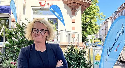 Eine lächelnde Frau mit blonden, lockigen Haaren und schwarzer Brille steht mit verschränkten Armen vor einem Reisebüro der TUI. Im Hintergrund sind blaue Werbeflaggen mit den Aufschriften „Robinson“ und „Mein Schiff – TUI Cruises“ zu sehen. Die Szene spielt an einem sonnigen Tag in einer belebten Stadtstraße mit Bäumen und Altbauten.