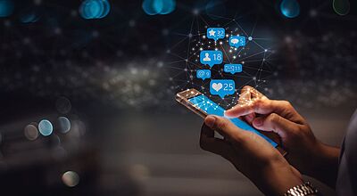 Eine Person hält ein Smartphone in der Hand und tippt auf den Bildschirm. Über dem Gerät schweben digitale Symbole für soziale Interaktionen, darunter Likes (Herzen), Nachrichten, Follower, E-Mails und Kommentare – jeweils mit Zahlen versehen. Die Darstellung ist futuristisch und mit leuchtenden, vernetzten Punkten auf dunklem Hintergrund versehen, was auf digitale Kommunikation und Social-Media-Aktivität hinweist.