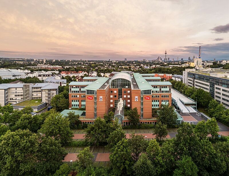 Luftaufnahme eines modernen Bürogebäudes mit Glasdach und roten Backsteinfassaden, umgeben von grünen Bäumen. Im Vordergrund verläuft ein Gehweg, im Hintergrund ist die Skyline von Frankfurt mit Fernsehturm und weiteren Hochhäusern bei Sonnenuntergang zu sehen. Das Gebäude trägt das Logo der DERTOUR Group.