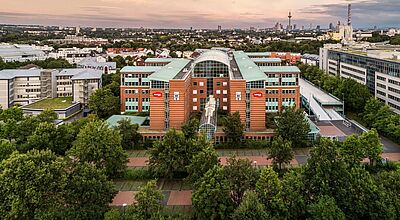 Die Dertour Group, hier die Zentrale in Frankfurt am Main, blickt Vorstellung der Sommerprogramme 2026 optimistisch in die neue Urlaubssaison Luftaufnahme eines modernen Bürogebäudes mit Glasdach und roten Backsteinfassaden, umgeben von grünen Bäumen. Im Vordergrund verläuft ein Gehweg, im Hintergrund ist die Skyline von Frankfurt mit Fernsehturm und weiteren Hochhäusern bei Sonnenuntergang zu sehen. Das Gebäude trägt das Logo der DERTOUR Group.