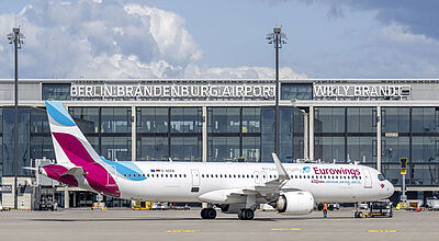 Ein Flugzeug der Gesellschaft Eurowings steht vor dem Terminalgebäude des Flughafens Berlin Brandenburg an einem sonnigen Tag mit blauem Himmel und Wolken.