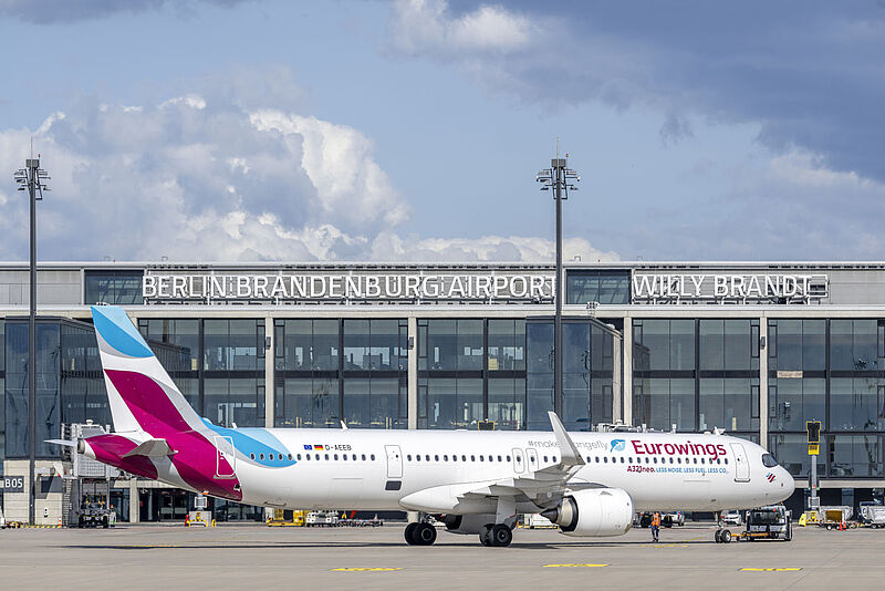 Ein Flugzeug der Gesellschaft Eurowings steht vor dem Terminalgebäude des Flughafens Berlin Brandenburg an einem sonnigen Tag mit blauem Himmel und Wolken.