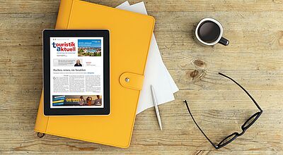 Tablet mit geöffneter digitalen Ausgabe der Zeitschrift touristik aktuell liegt auf einem gelben Ordner auf einem Holztisch, daneben eine Tasse Kaffee, ein Kugelschreiber, ein paar Blätter Papier und eine schwarze Brille.