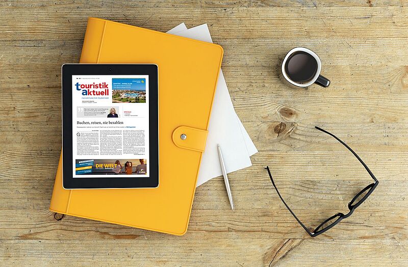 Tablet mit geöffneter digitalen Ausgabe der Zeitschrift touristik aktuell liegt auf einem gelben Ordner auf einem Holztisch, daneben eine Tasse Kaffee, ein Kugelschreiber, ein paar Blätter Papier und eine schwarze Brille.