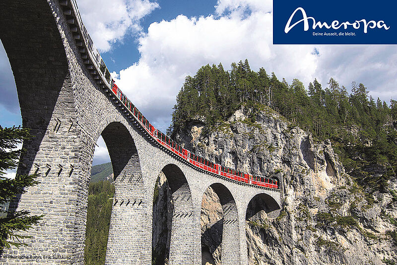 Panoramaaufnahme der berühmten Landwasser Viadukt-Brücke in der Schweiz, mit einem roten Zug der Rhätischen Bahn, der die beeindruckende Brücke überquert. Die Szene zeigt eine wunderschöne Berglandschaft mit grünen Wäldern und einem klaren blauen Himmel, perfekt für eine unvergessliche Zugreise durch die Schweizer Alpen