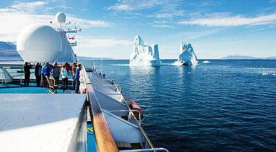 Der neue Name steht für das erweiterte Arktis-Programm des Island-Spezialisten Eine Gruppe Kreuzfahrttouristen steht an Deck eines Expeditionsschiffes und blickt auf zwei aus dem Wasser aufragende Eisberge.