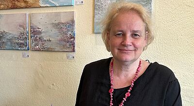 Eine Frau mit blondem Haar, schwarzer Bluse und pinkfarbener Halskette hält eine Auszeichnung aus Glas in der Hand. Die Auszeichnung trägt den Namen Sylvia Raschke und stammt von der Central America Tourism Agency (CATA). Im Hintergrund hängen mehrere Gemälde mit Meeres- und Küstenmotiven an einer hellen Wand.