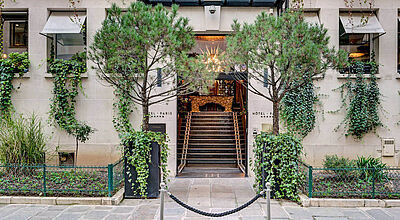 Eingang eines eleganten Boutique-Hotels in Paris, flankiert von zwei Bäumen in Pflanzgefäßen. Die Fassade ist mit hellen Steinen verkleidet und von grünen Kletterpflanzen bewachsen. Eine Treppe führt unter einem modernen Glasvordach zum Haupteingang, durch den ein kunstvoller Kronleuchter sichtbar ist