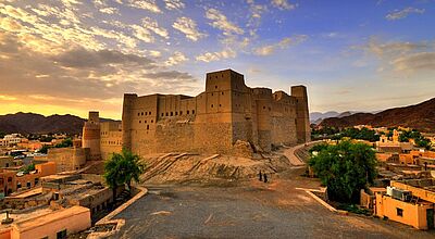 „Mächtige historische Festung in Oman bei Sonnenuntergang, umgeben von kleinen Häusern, Bergen im Hintergrund und einem dramatischen Himmel mit Wolken.“