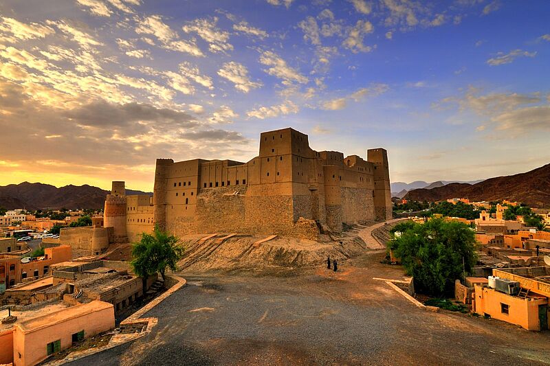 „Mächtige historische Festung in Oman bei Sonnenuntergang, umgeben von kleinen Häusern, Bergen im Hintergrund und einem dramatischen Himmel mit Wolken.“