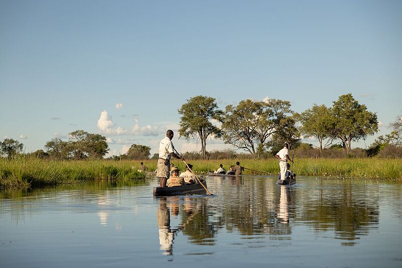 Reisende in traditionellen Holzkanus mit lokalen Guides fahren durch ein Flussdelta in Botswana, umgeben von Graslandschaft und Bäumen unter blauem Himmel.