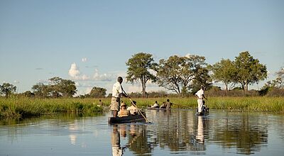 Reisende in traditionellen Holzkanus mit lokalen Guides fahren durch ein Flussdelta in Botswana, umgeben von Graslandschaft und Bäumen unter blauem Himmel.