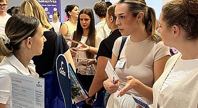 Junge Touristikerinnen im Gespräch an einem Messestand von Costa Kreuzfahrten beim Nachwuchs-Camp von Best-Reisen; auf dem Tisch liegen Broschüren, Kugelschreiber, Lanyards und Infomaterial zur Beratung am Counter. Im Hintergrund weitere Teilnehmende im Austausch.
