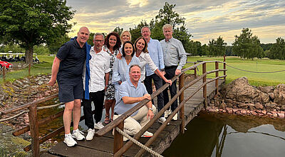 Eine Gruppe von neun Erwachsenen steht auf einer kleinen Holzbrücke über einem Teich auf einem Golfplatz. Im Hintergrund sind grüne Wiesen, Bäume und Golfwagen zu sehen. Der Himmel ist bewölkt mit einer aufklarenden Abendstimmung. Die Personen lachen und posieren fröhlich für das Foto, einige sind leger gekleidet, andere schicker.